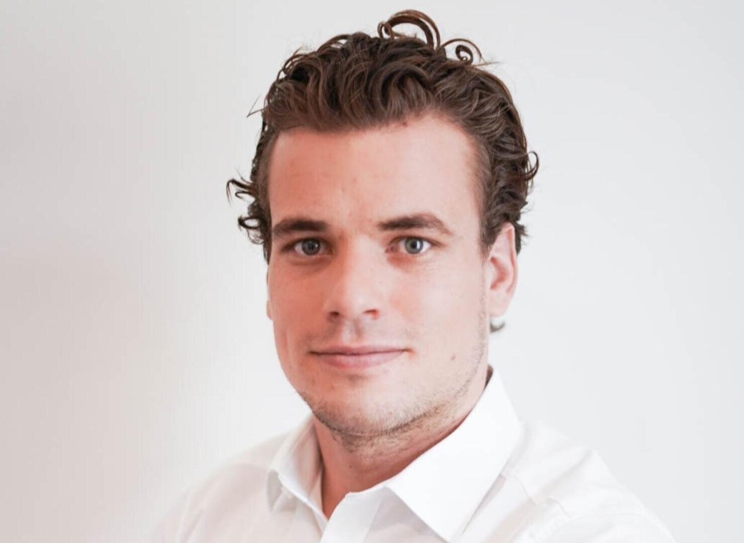 Ein Mann mit kurzen, lockigen braunen Haaren und blauen Augen, der ein weißes Hemd mit Kragen trägt, posiert vor einem schlichten weißen Hintergrund und schaut mit einem leichten Lächeln direkt in die Kamera.