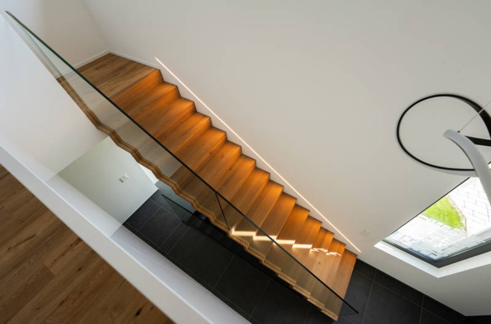 Eine moderne Treppe mit Holzstufen und Glasgeländer von oben betrachtet. Die Treppe ist mit warmem Licht beleuchtet, der Boden darunter ist dunkelgrau gefliest. Ein großes Fenster lässt Tageslicht herein.