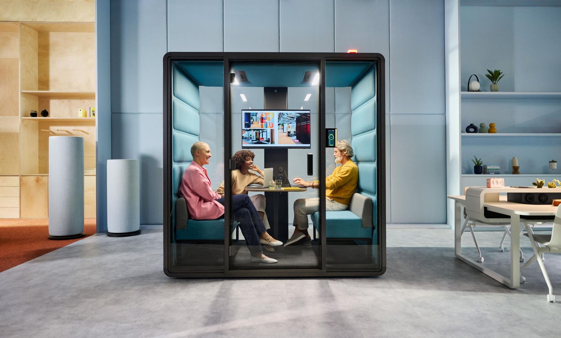 Trois personnes sont assises dans une cellule de bureau vitrée et tiennent une réunion. Ils se font face autour d'une table avec des ordinateurs portables et des notebooks, tandis qu'à l'extérieur de la capsule, on aperçoit des étagères colorées et un espace de bureau moderne.