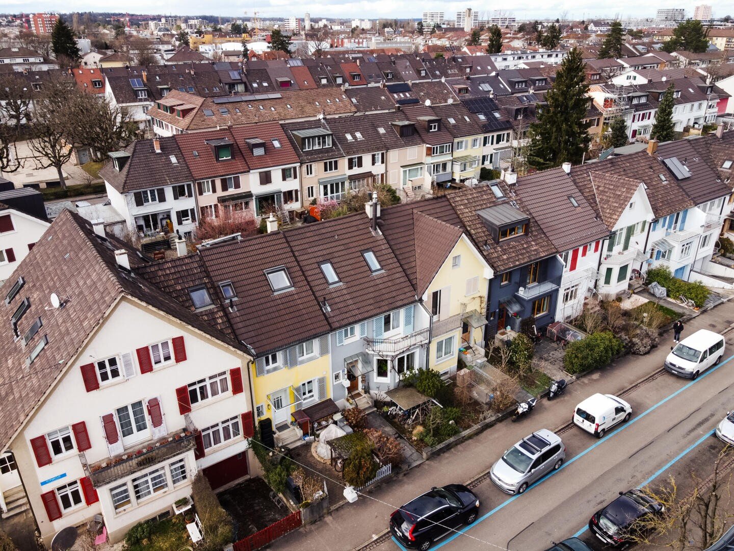 Luftaufnahme einer mit bunten Reihenhäusern, geparkten Autos und kleinen Vorgärten gesäumten Wohnstraße. Die Nachbarschaft wirkt ruhig, mit mehreren Reihen ähnlicher Häuser und modernen Gebäuden im Hintergrund.