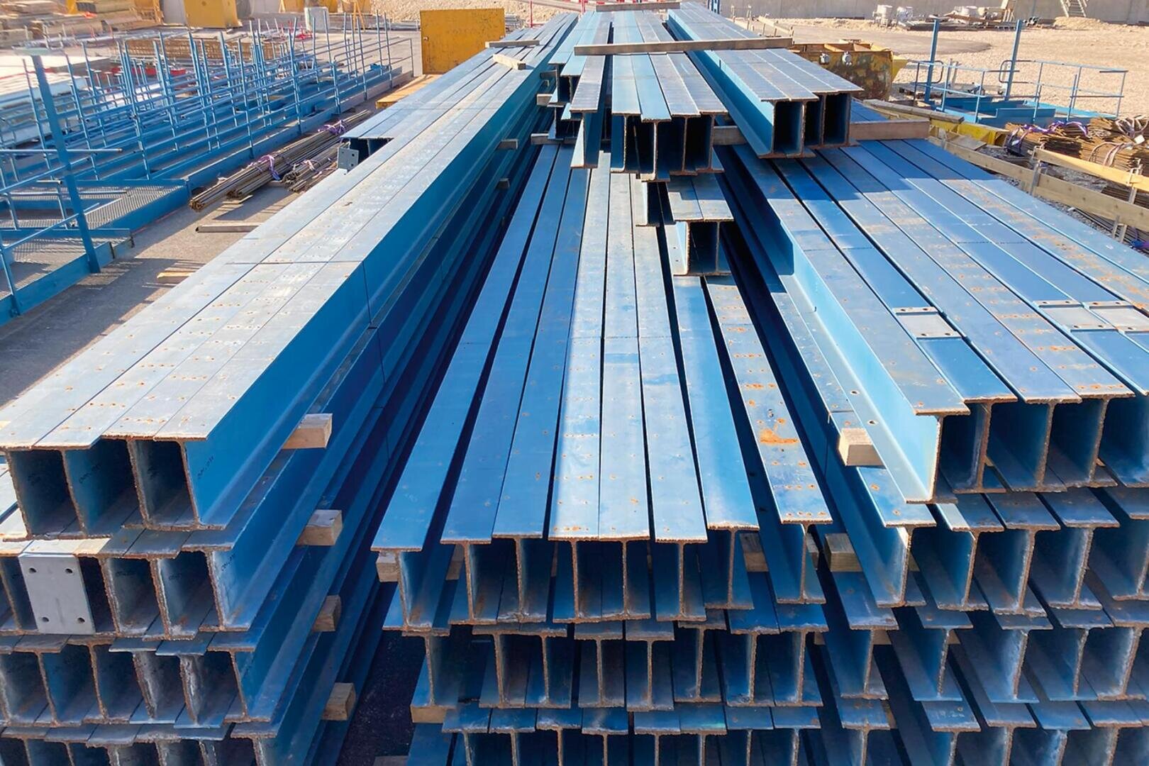 Stapel von langen, blau gestrichenen Stahlträgern sind auf einer Baustelle ordentlich angeordnet, während im Hintergrund andere Materialien und Geräte zu sehen sind.
