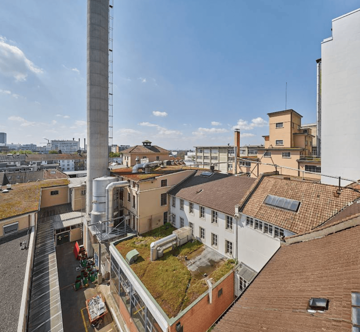 Ein Blick auf einen Industriekomplex mit hellbraunen Gebäuden, einem hohen Schornstein, Dachbegrünung und verschiedenen Geräten. Der Himmel ist klar mit ein paar Wolken, und im Hintergrund sind städtische Gebäude zu sehen.