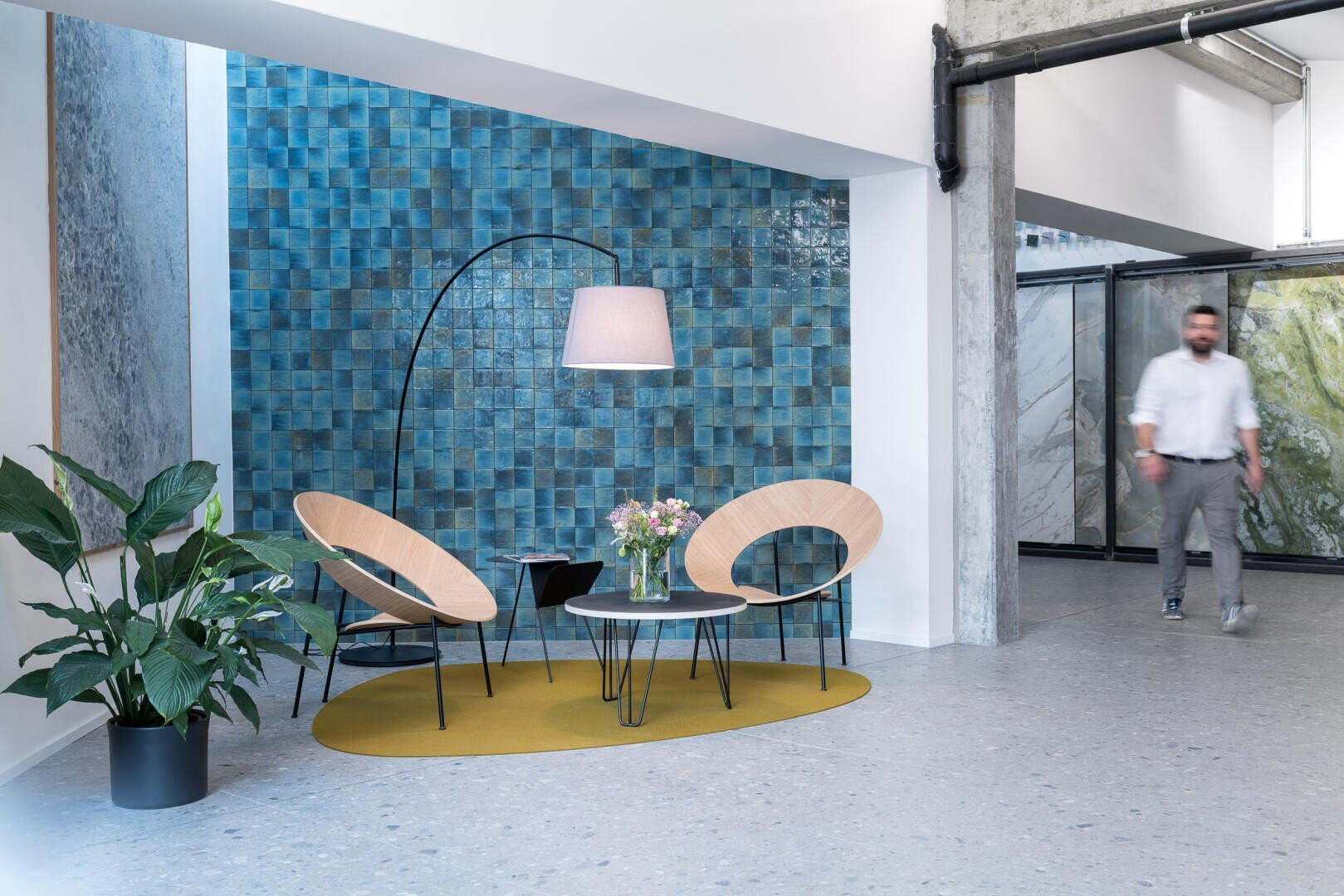 Salon de bureau moderne avec deux chaises rondes en bois, une petite table ronde avec un bouquet de fleurs, un lampadaire et une plante en pot. Un mur d'accent carrelé bleu et une personne qui marche en arrière-plan.