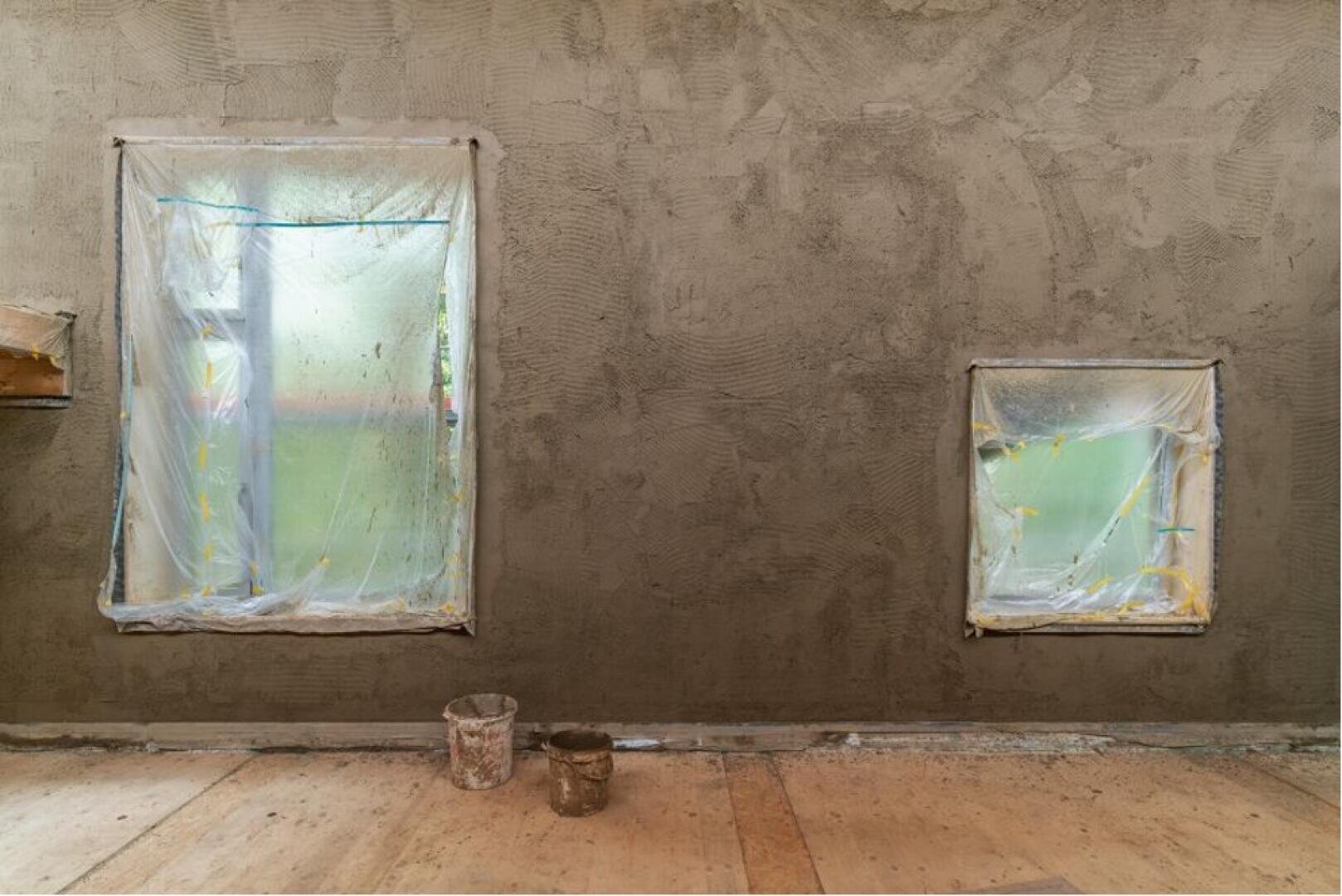 Zwei mit Plastikplanen und Klebeband abgedeckte Fenster sind in eine frisch verputzte Wand eingelassen. Zwei Eimer stehen auf einem nackten Holzboden, was auf laufende Renovierungs- oder Bauarbeiten hindeutet.