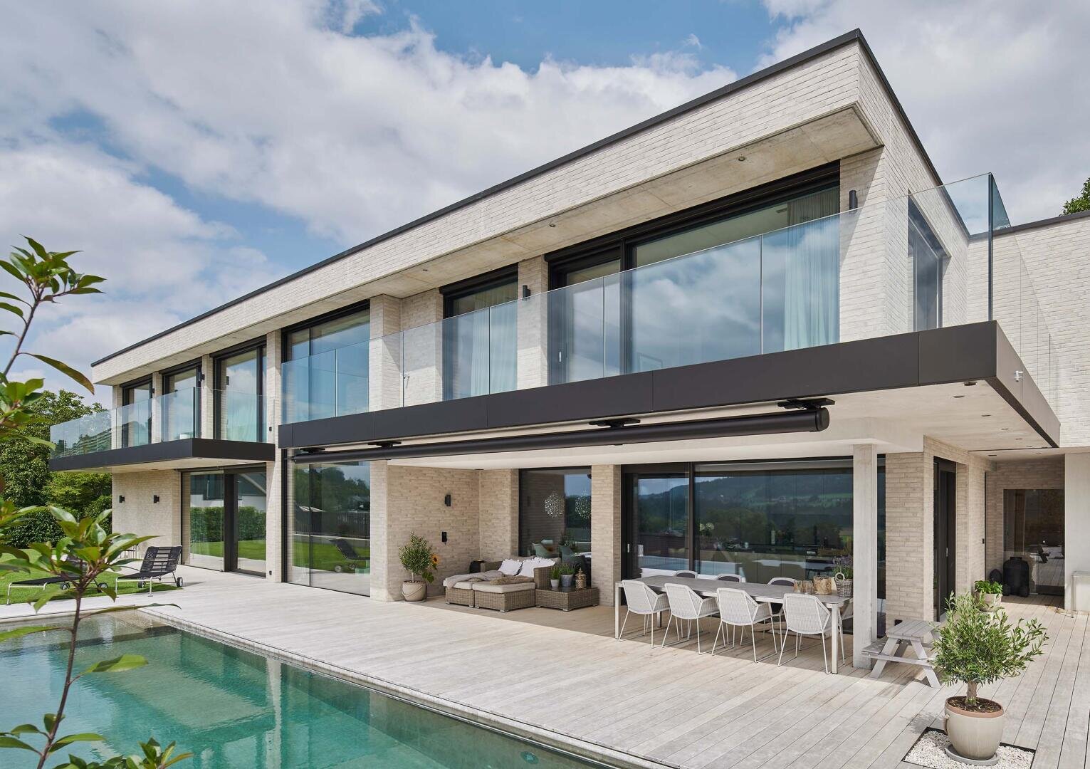 Une maison moderne de deux étages avec de grandes baies vitrées et des balcons, une salle à manger en plein air, des sièges et une piscine, entourée de verdure sous un ciel partiellement nuageux.
