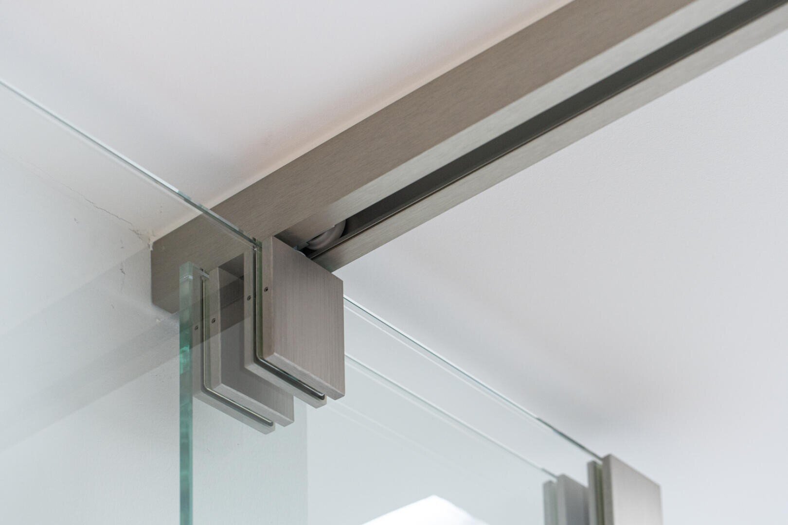 Nahaufnahme einer modernen Glasschiebetür mit einer Metallschiene und einer an der Decke montierten Halterung, die die Beschläge zur Befestigung der Glasscheibe an der Schiene zeigt.