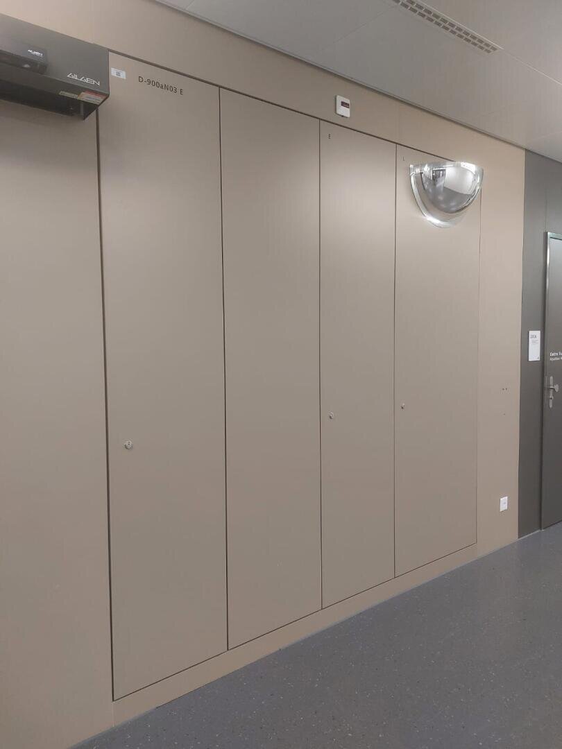 Eine beigefarbene Wand mit hohen, schmalen, bündig abschließenden Türen, eine kuppelförmige Leuchte, eine kleine Überwachungskamera und ein Lüftungsschacht für eine Klimaanlage in einem sauberen Flur mit grau gesprenkeltem Boden.