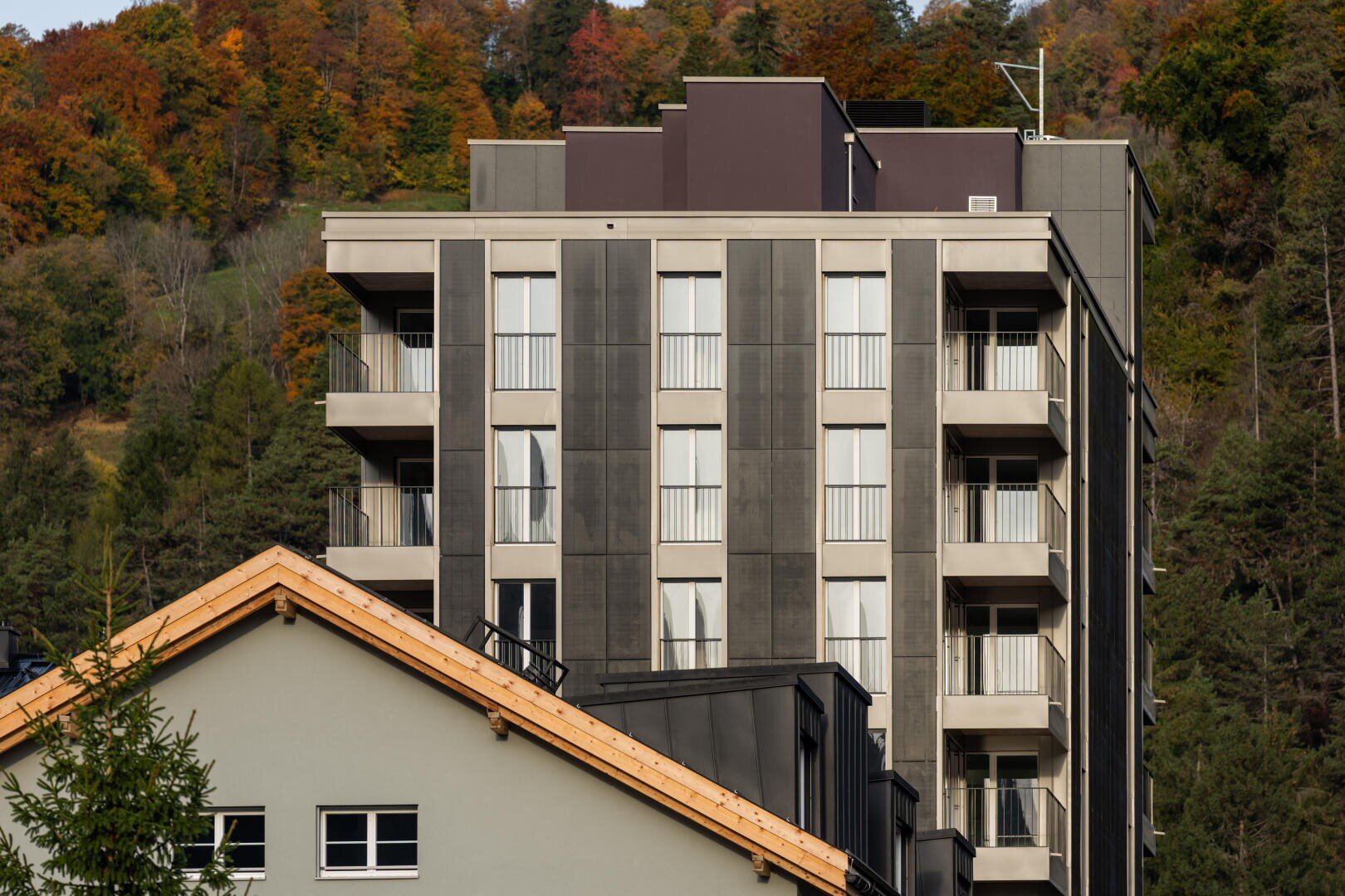 Ein modernes Mehrfamilienhaus mit Balkonen steht vor einem beigen Haus mit Holzdach, das sich vor dem Hintergrund von Bäumen mit Herbstlaub an einem Hang befindet.