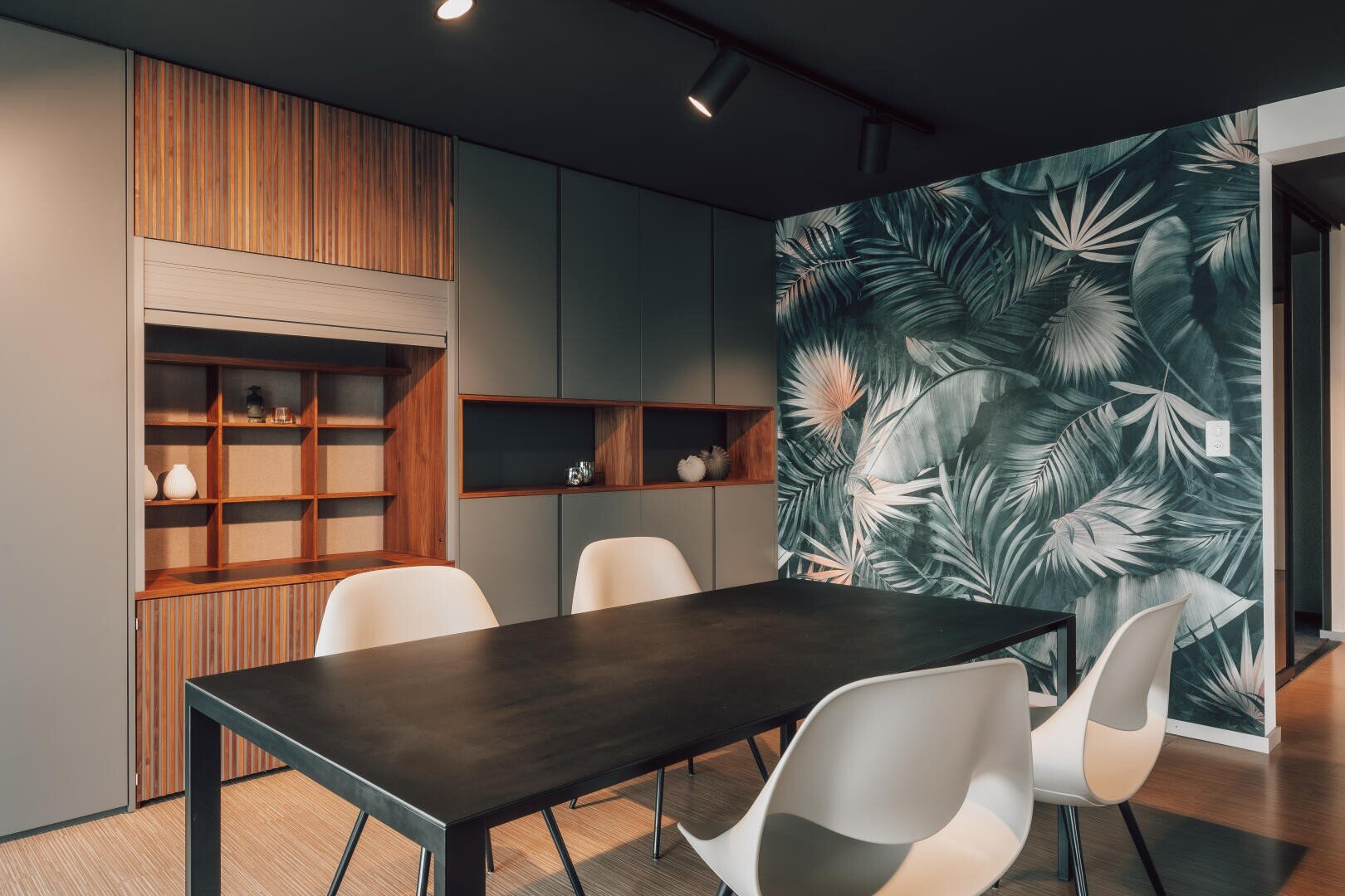 Ein modernes Esszimmer mit einem schwarzen Tisch, vier weißen Stühlen, Einbauschränken aus Holz und Grau und einer Wand, die mit einem kühnen Wandbild mit grünen und grauen tropischen Blättern verziert ist.