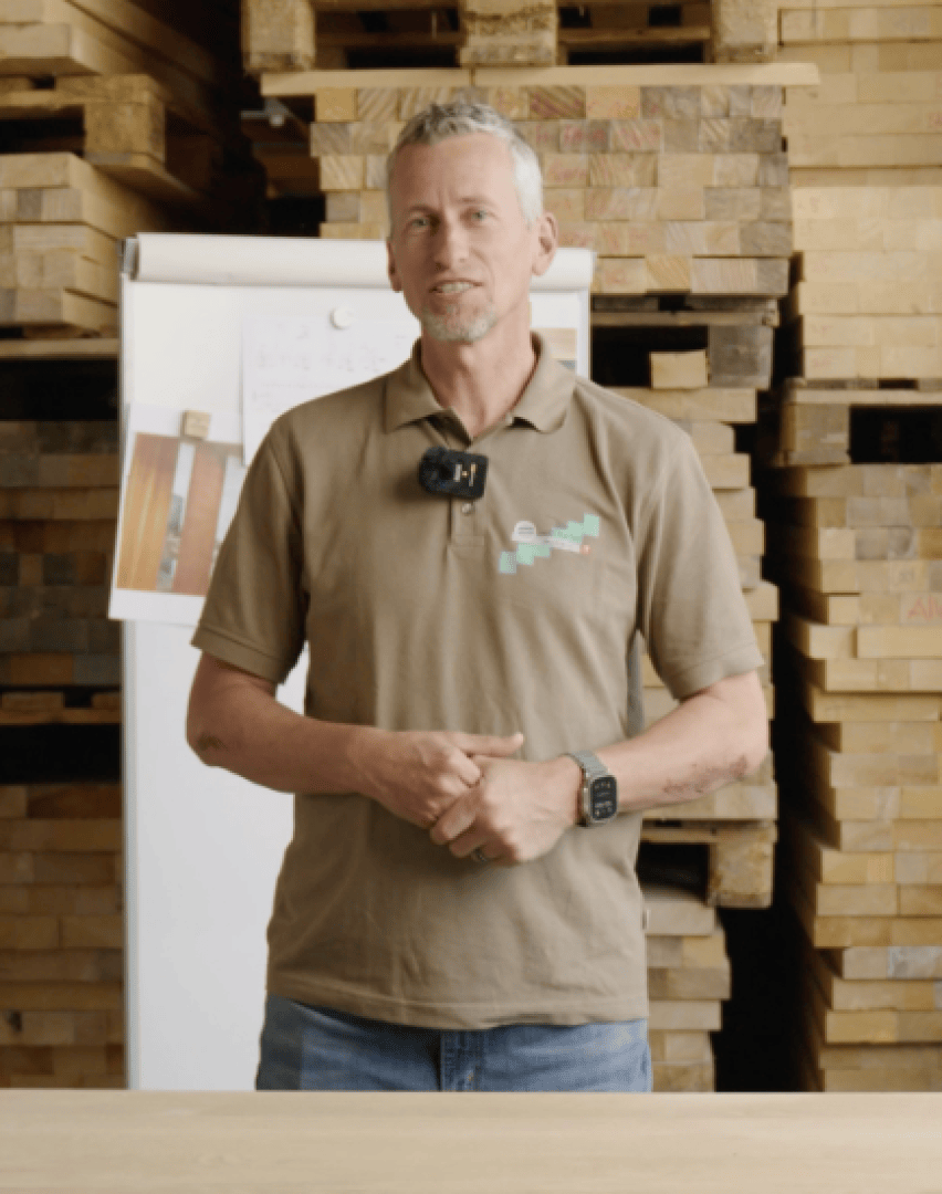 Ein Mann in einem braunen Poloshirt steht vor gestapelten Holzbrettern. Er hat kurze graue Haare, einen Bart und trägt eine Smartwatch und ein Mikrofon. Im Hintergrund ist ein Whiteboard zu sehen.