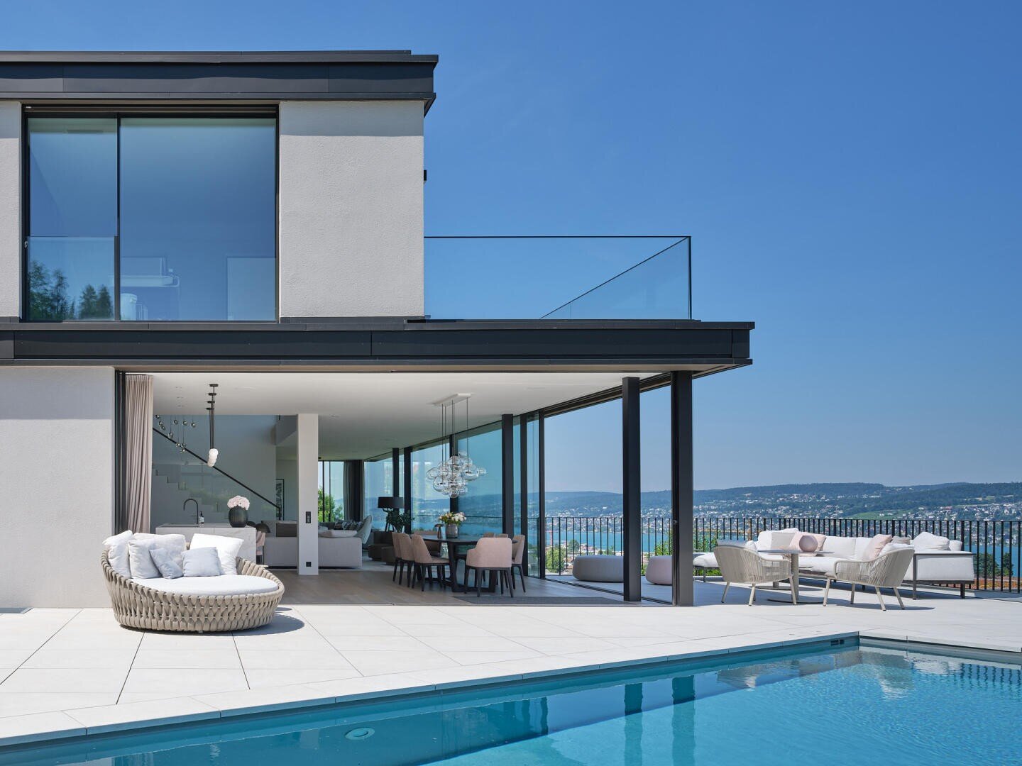 Maison moderne de deux étages avec de grandes baies vitrées, une piscine extérieure, des chaises longues et un coin repas sur la terrasse donnant sur un paysage pittoresque de collines et une ville sous un ciel bleu.