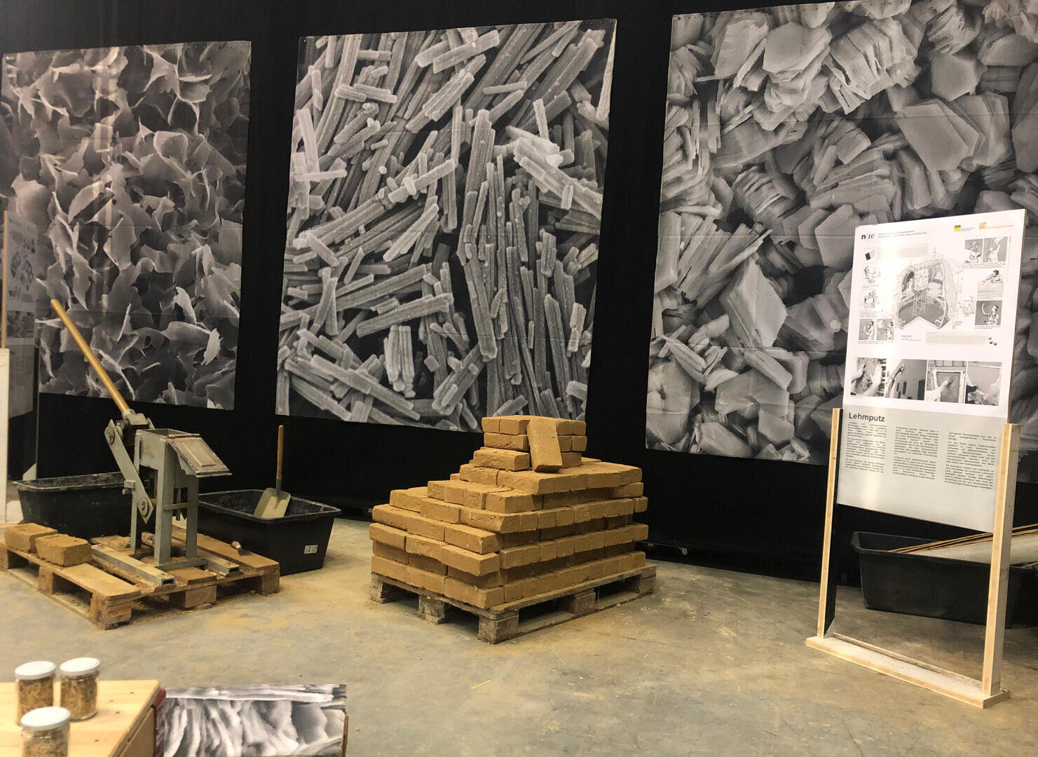 Ausstellungsdisplay mit großen mikroskopischen Graustufenbildern, einem Stapel rechteckiger Ziegelsteine auf einer Holzpalette, Bauwerkzeugen, Informationspostern und Baumaterialien auf einem Betonboden.