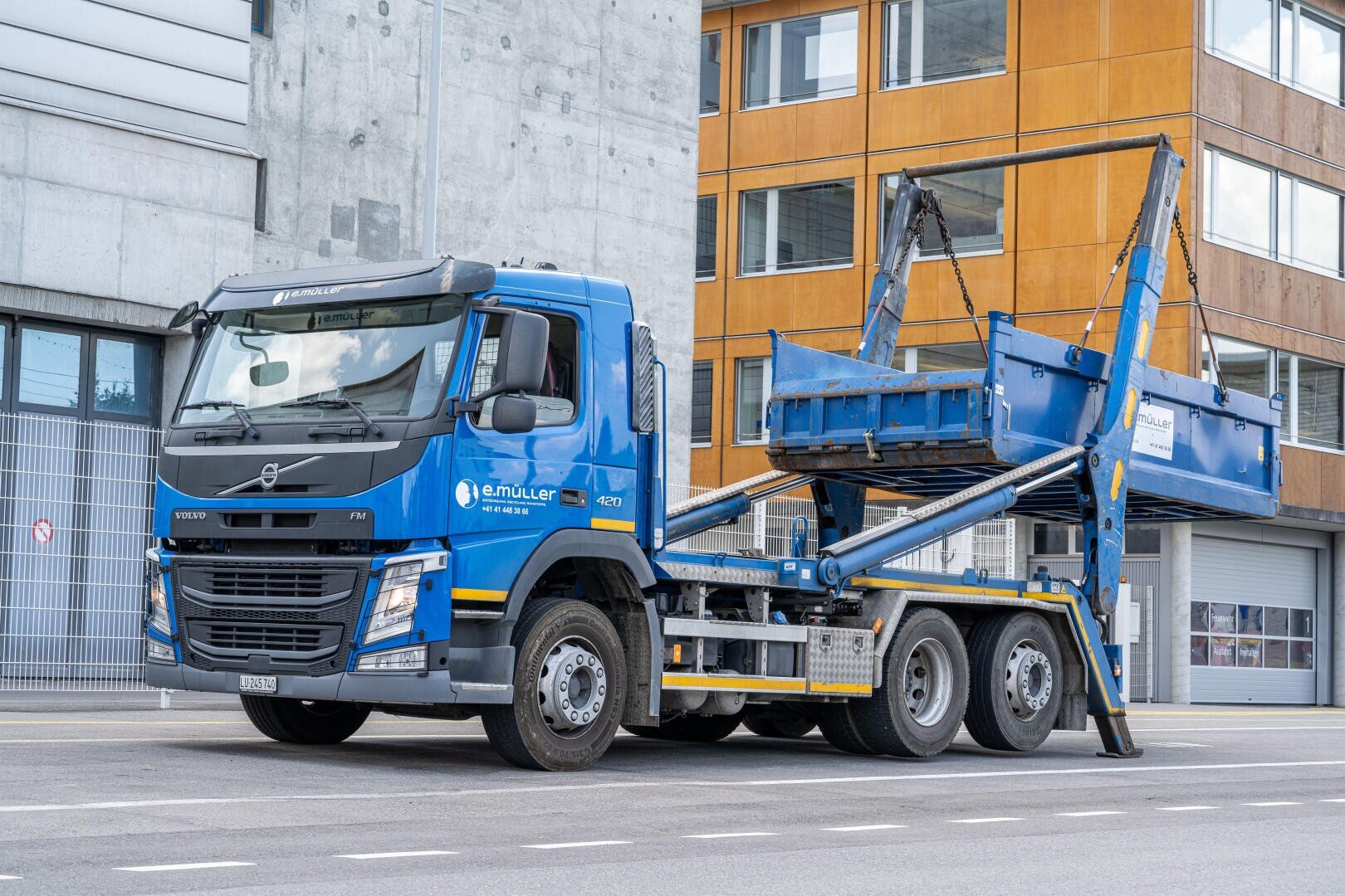 Ein blauer Volvo-Lkw mit einem großen Metallcontaineraufzug ist auf einer Straße in der Stadt neben einem modernen Gebäude mit orangefarbenen und grauen Wänden geparkt. Der Container auf dem Lkw ist in eine aufrechte Position gehoben.