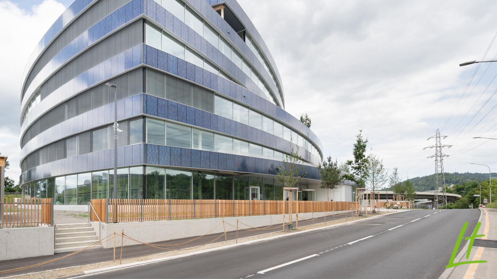 Ein modernes, geschwungenes Glasgebäude mit blauen und grauen Paneelen steht neben einer leeren, von Bäumen und Holzzäunen gesäumten Straße unter einem bewölkten Himmel. Im Hintergrund sind Stromleitungen und Hügel zu sehen.