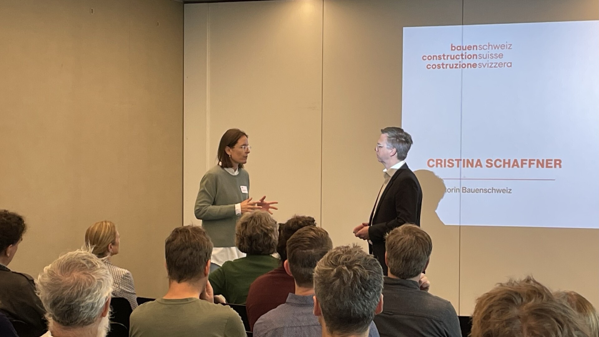 Zwei Personen stehen während einer Präsentation vor einem sitzenden Publikum und unterhalten sich. Eine Folie im Hintergrund zeigt den Namen Cristina Schaffner und das Logo von bauenschweiz.