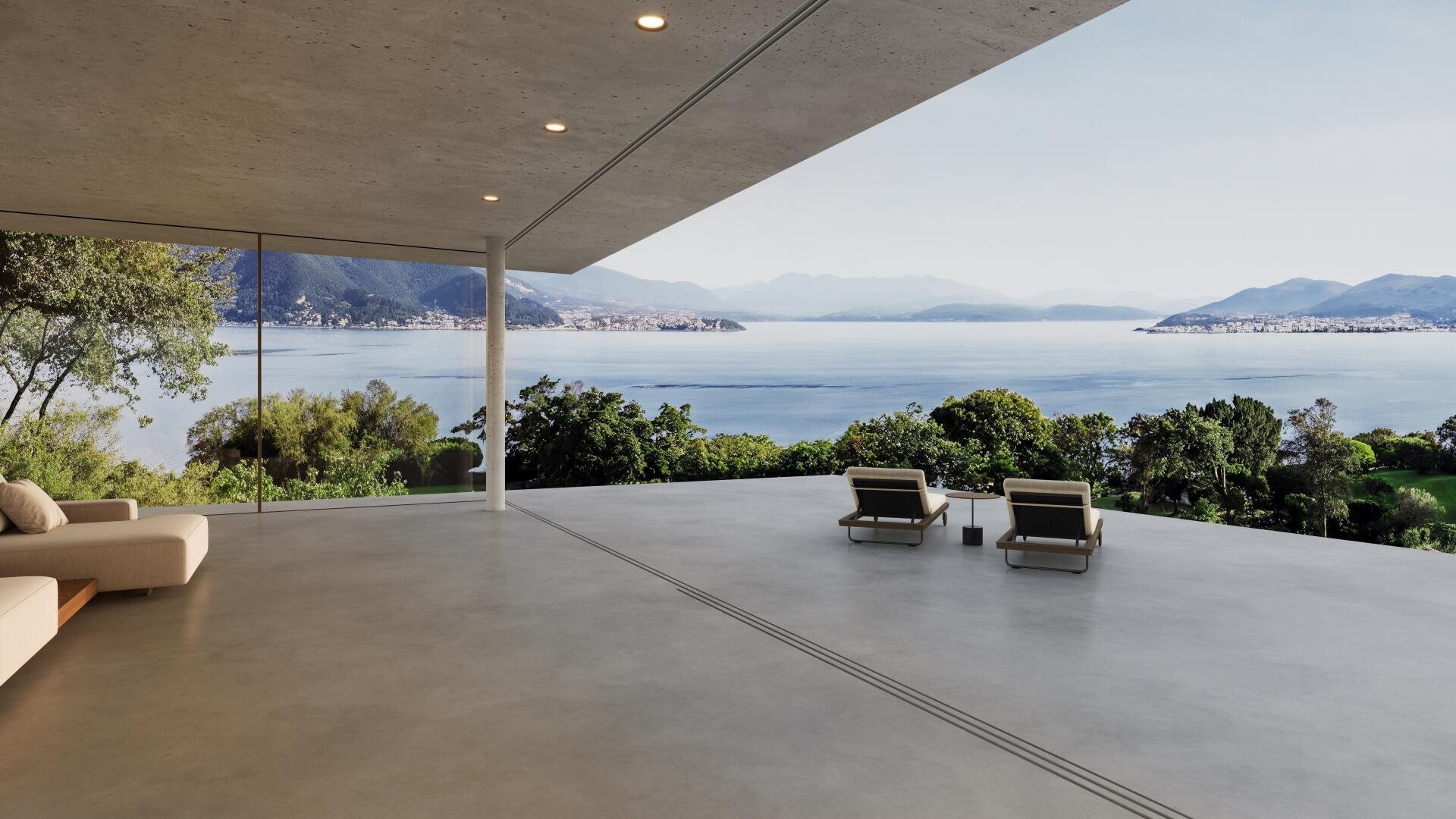 Eine geräumige, moderne Terrasse mit zwei Loungesesseln überblickt einen malerischen See und die fernen Berge, umrahmt von Grünflächen und großen Glaswänden, unter einem klaren Himmel.