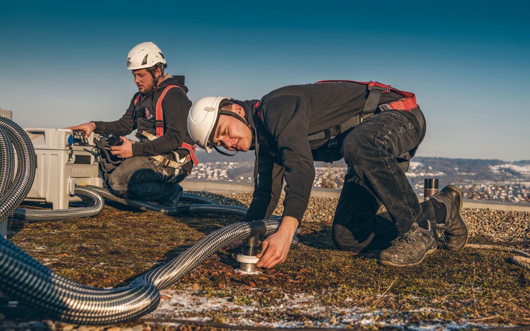 Zwei Arbeiter in Schutzkleidung und mit Helmen installieren Geräte auf einem grünen Dach. Einer kniet, um ein Rohr einzustellen, während der andere ein Steuergerät bedient. Um sie herum sind Rohre und Kabel verlegt, im Hintergrund ist eine Stadtlandschaft zu sehen.