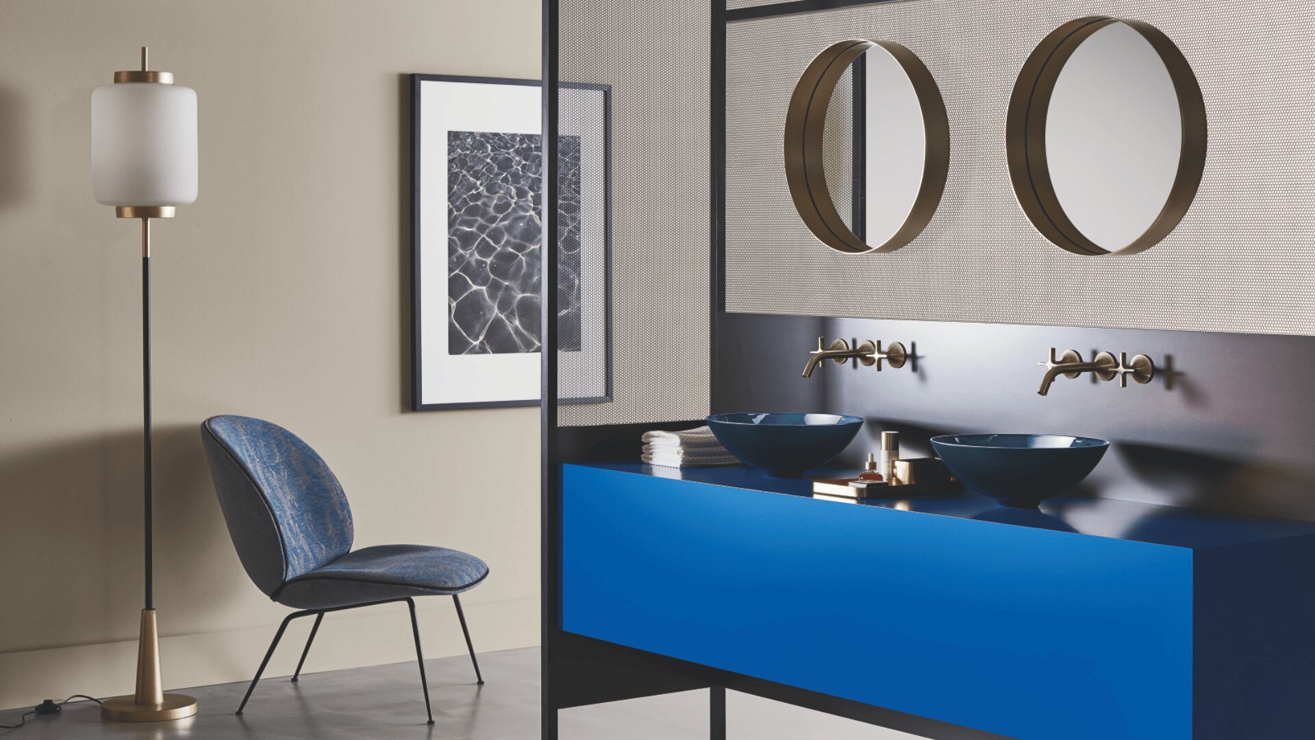 Ein modernes Badezimmer mit einem blauen Waschtisch, zwei runden Spiegeln, Gefäßwaschbecken, wandmontierten Armaturen, einer Stehlampe und einem blauen Stuhl. An der beigen Wand hängt ein gerahmtes Schwarz-Weiß-Foto.