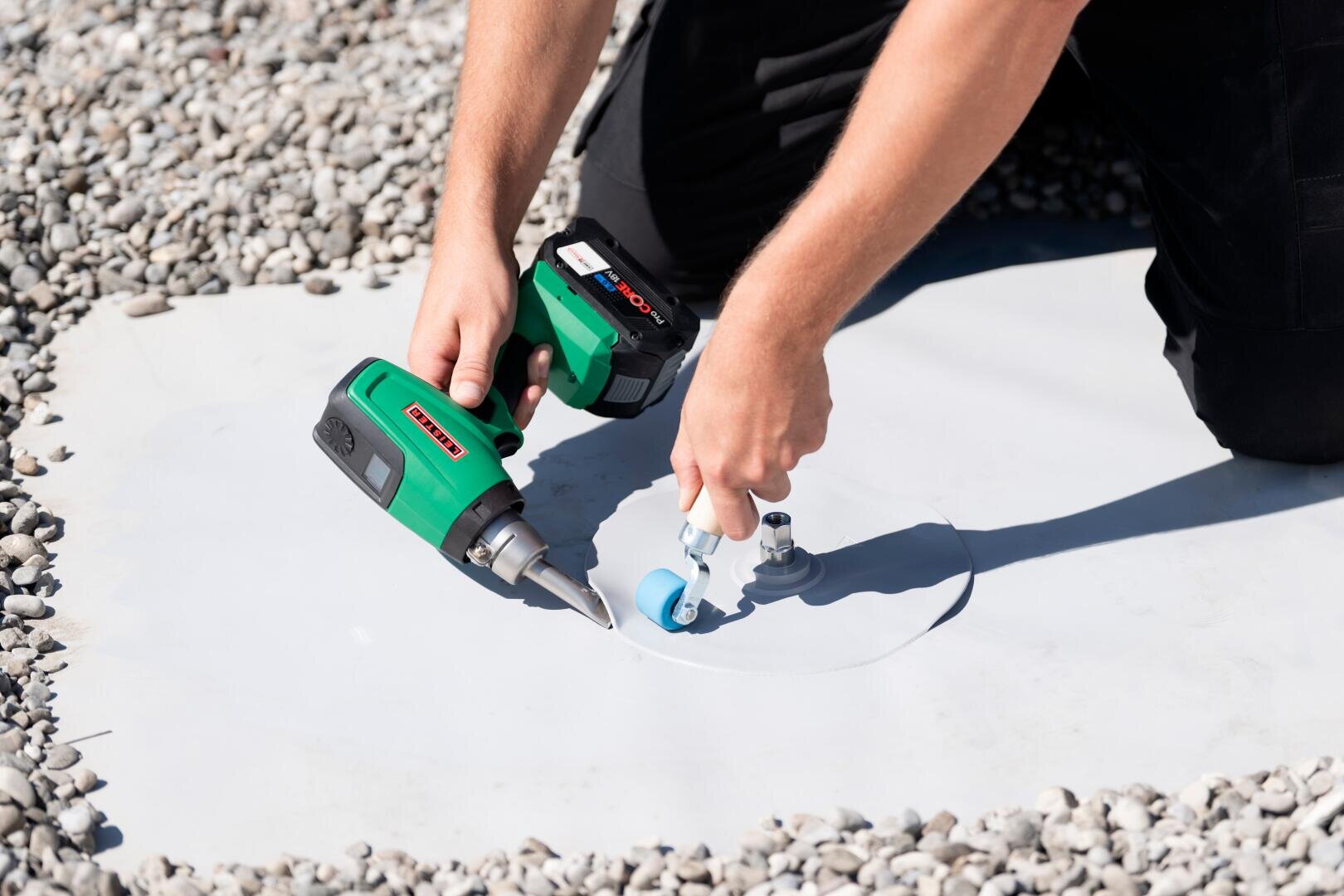 Une personne travaille avec une perceuse sans fil verte et un outil à ventouse sur une surface blanche à l'extérieur, entourée de petits cailloux gris. Seuls les bras et les mains de la personne sont visibles.