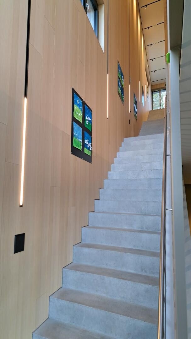 Eine moderne Innentreppe mit Betonstufen und einem Metallhandlauf, flankiert von einer hohen Holzwand mit bunten Kunstwerken und vertikalen Lichtbändern. Durch die hohen Fenster strömt natürliches Licht herein.