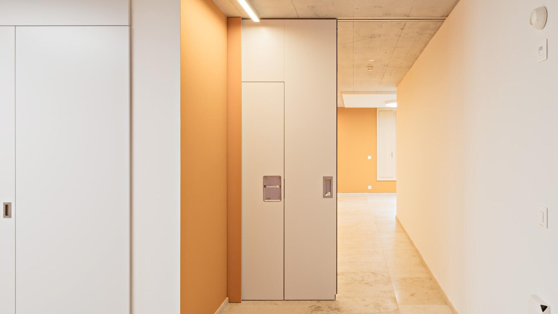 Ein moderner, minimalistischer Flur mit beigen Bodenfliesen, orangefarbenen und weißen Wänden, Einbaubeleuchtung und eleganten weißen Schiebetüren. Das Design ist sauber und übersichtlich und schafft eine helle, offene Atmosphäre.