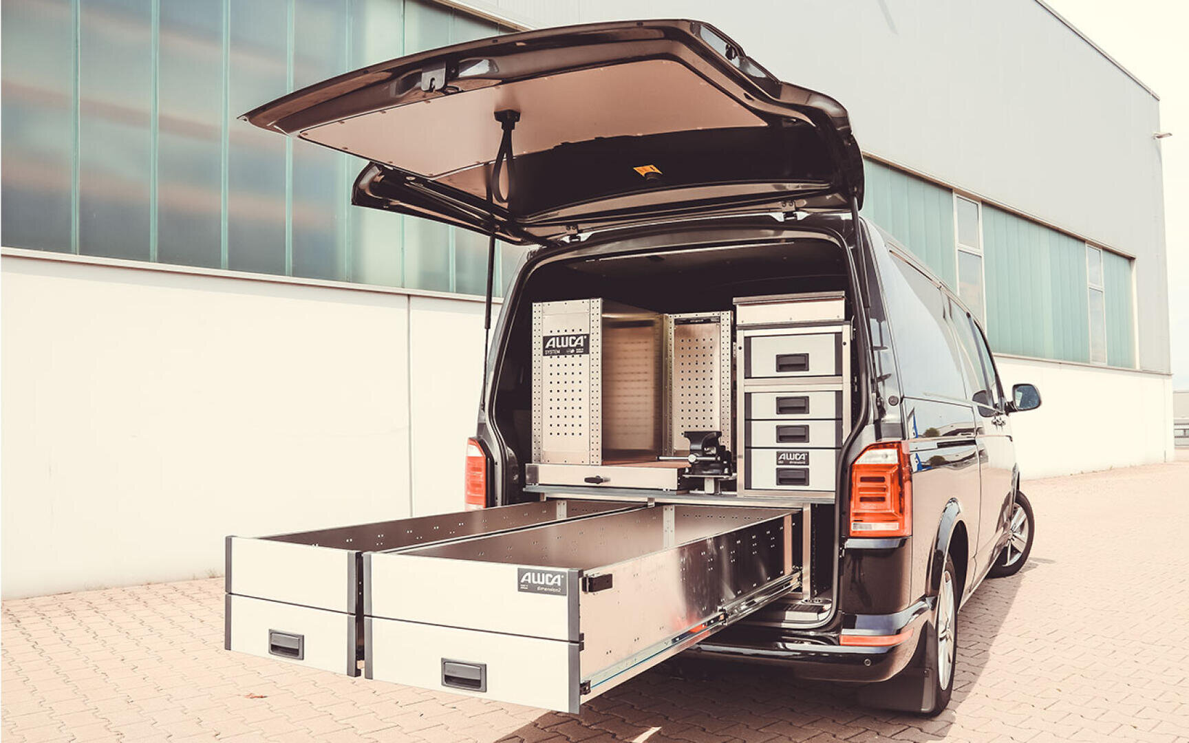 Ein schwarzer Lieferwagen mit geöffneten Hecktüren, die ein organisiertes Lagersystem mit Metallschubladen, Regalen und Fächern offenbaren, parkt vor einem modernen Industriegebäude.