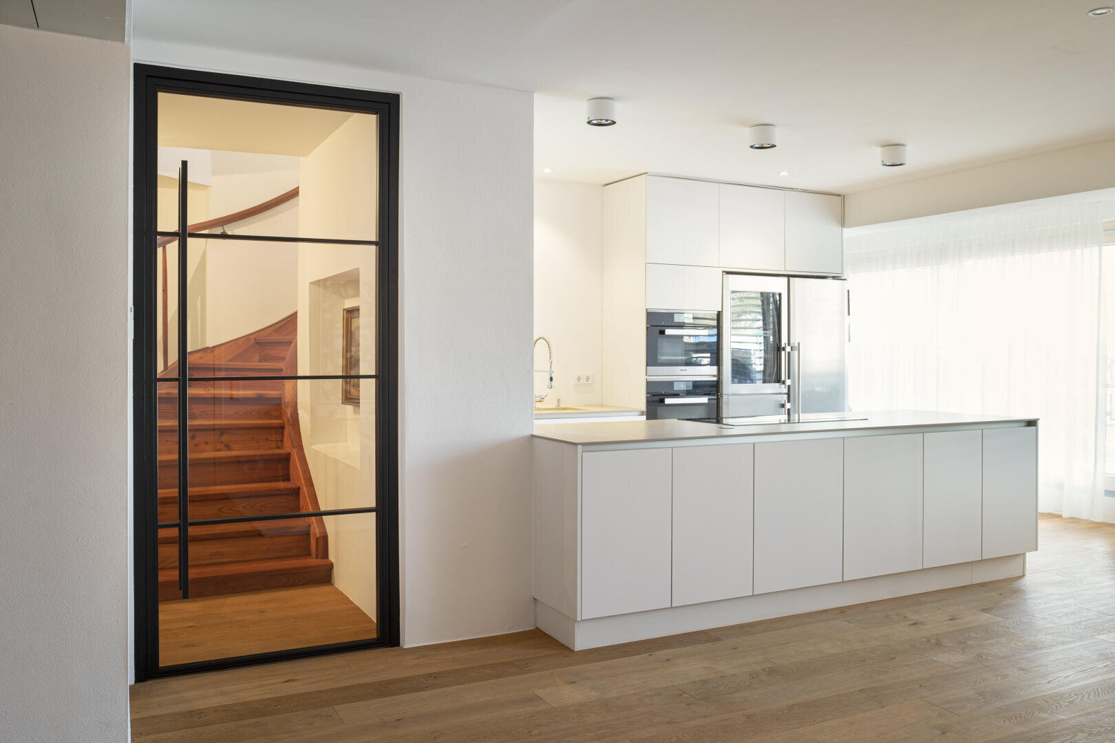 Eine moderne, minimalistische Küche mit weißen Schränken, Einbaugeräten, einer großen Insel, hellem Holzfußboden und einer Glastür, die eine Holztreppe offenbart. Natürliches Licht durchflutet den hellen, offenen Raum.