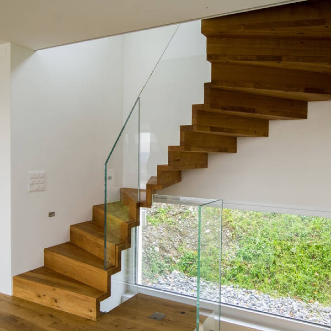 Un escalier moderne en bois avec contremarches ouvertes et rampes en verre monte dans une pièce lumineuse avec des murs blancs et une grande fenêtre donnant sur la verdure extérieure.