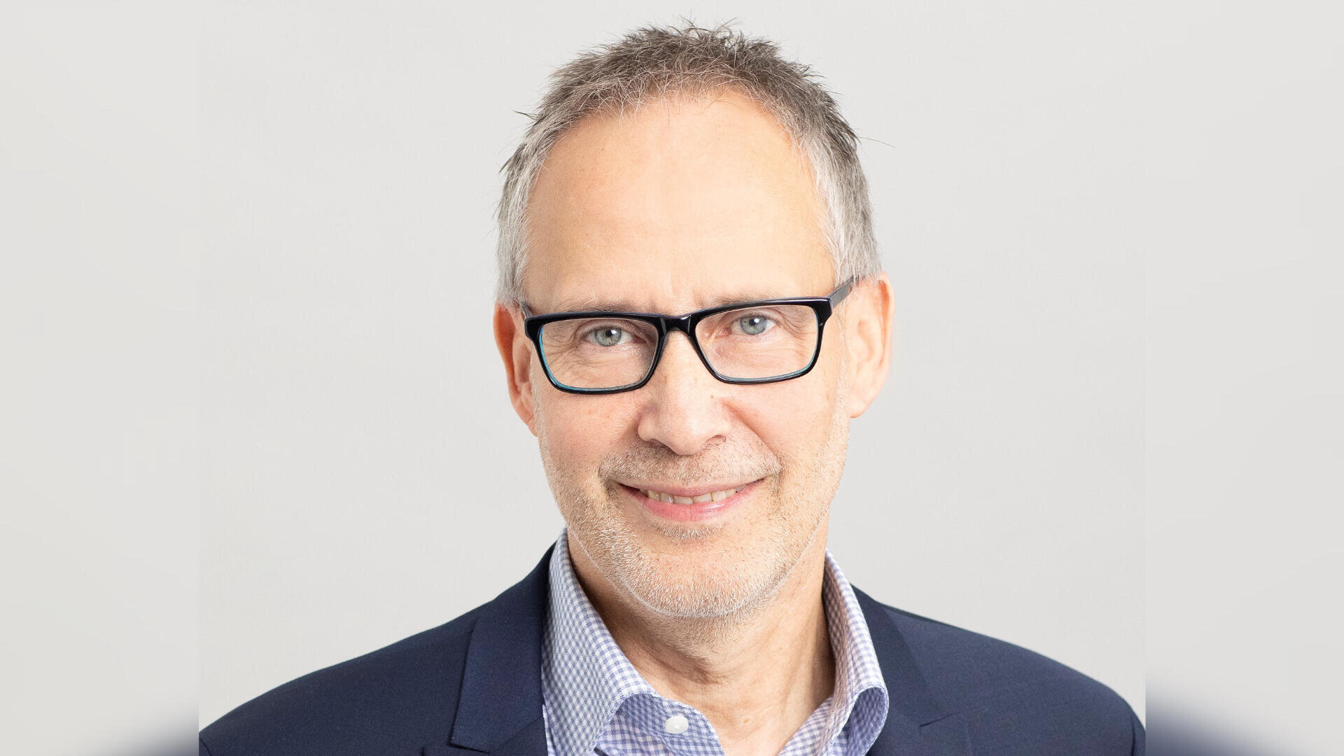 Ein Mann mittleren Alters mit kurzen grauen Haaren, einer schwarzen Brille, einem karierten Hemd und einem dunklen Blazer lächelt vor einem schlichten hellgrauen Hintergrund in die Kamera.