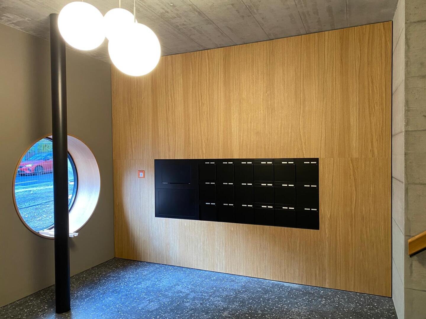 Ein modernes Interieur mit einer Holzwand mit schwarzen Briefkästen, einem runden Fenster auf der linken Seite, drei kugelförmigen Deckenleuchten und einer schwarzen Stange neben dem Fenster. Der Fußboden ist dunkel mit einem gesprenkelten Muster.