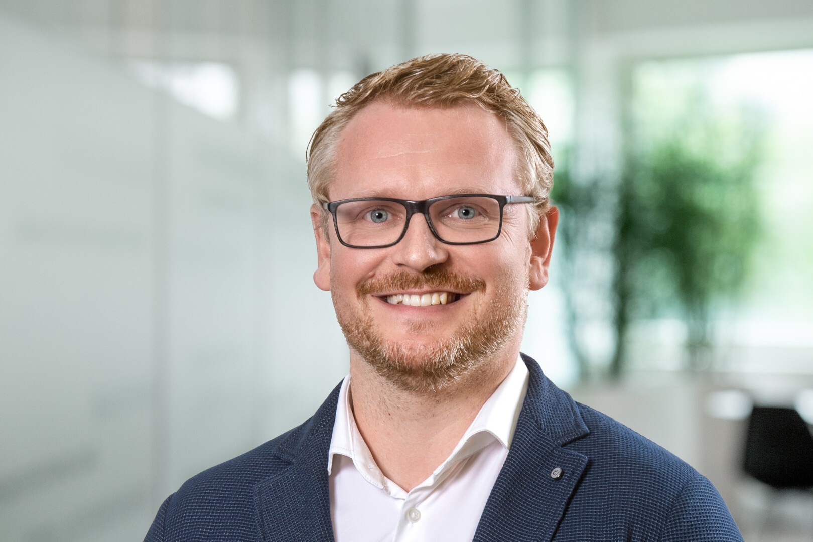 Ein lächelnder Mann mit blondem Haar und Bart, Brille, weißem Hemd und blauem Blazer steht in einem hellen Büro mit verschwommenen Fenstern und Grün im Hintergrund.