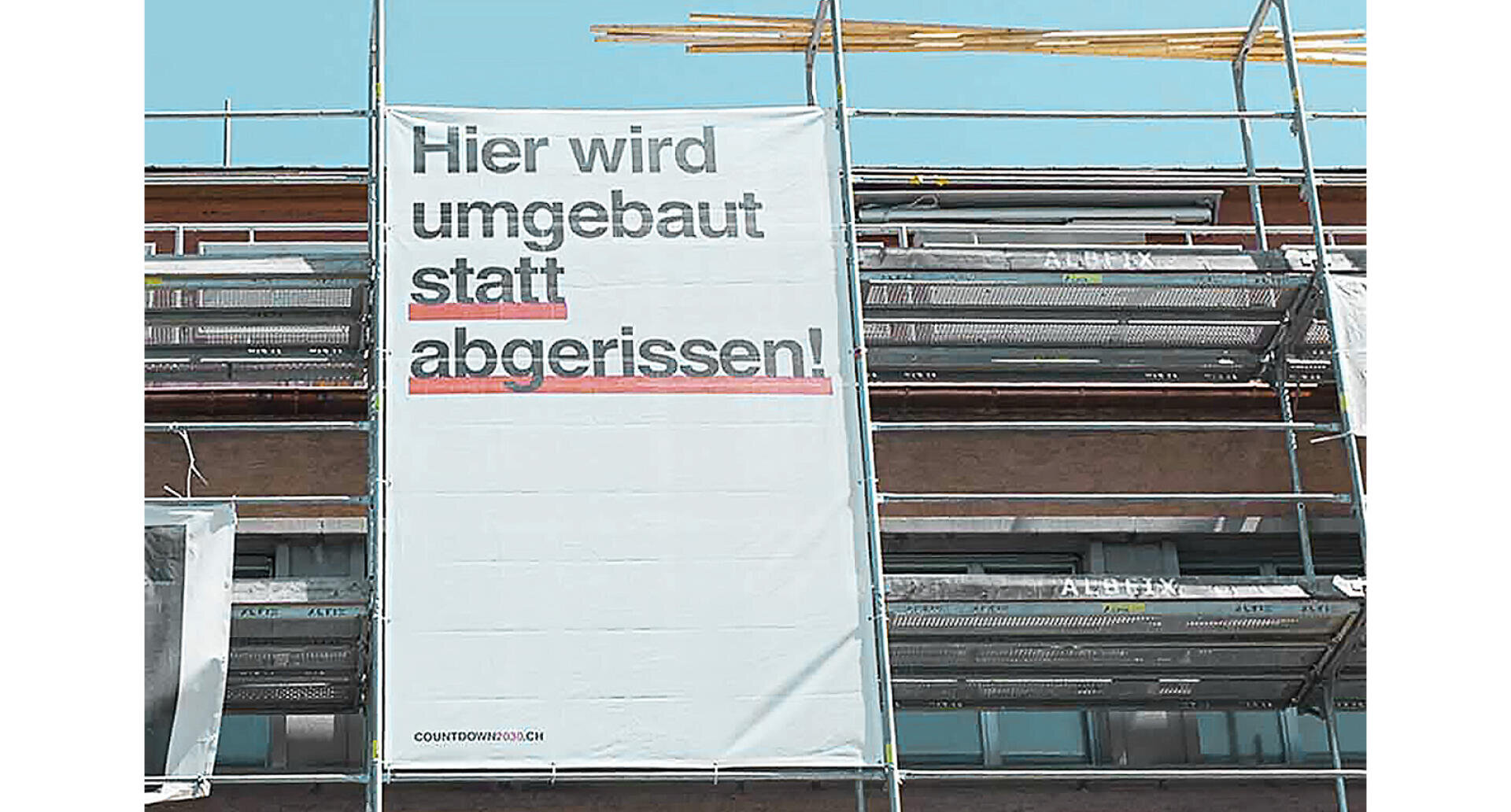 Auf einem großen weißen Banner auf einem Baugerüst steht "Hier wird umgebaut statt abgerissen!", was so viel bedeutet wie "Hier wird umgebaut statt abgerissen!" Baumaterialien und blauer Himmel sind zu sehen.