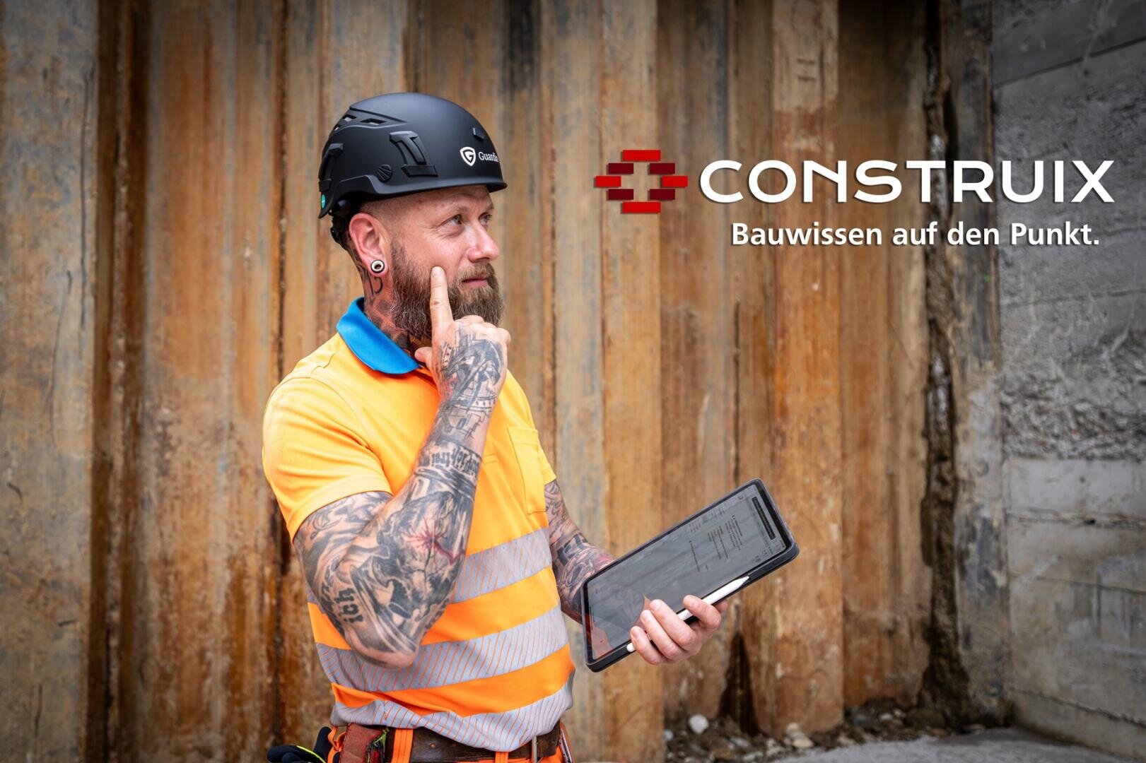 Ein Bauarbeiter mit Tattoos, der einen Helm und eine orangefarbene Sicherheitsweste trägt, hält ein Tablet und berührt nachdenklich sein Gesicht. Hinter ihm steht eine Holzwand, auf der rechts das Logo CONSTRUIX Bauwissen auf den Punkt.