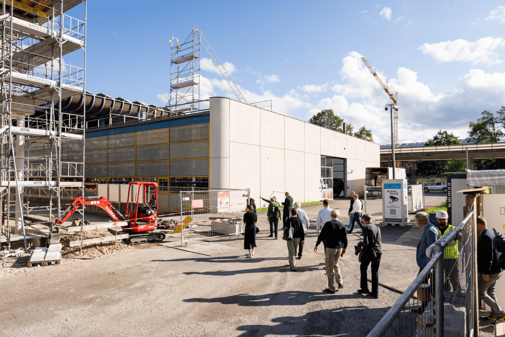 Eine Gruppe von Menschen geht an einer Baustelle mit Gerüsten, einem roten Bagger und einem modernen weißen Gebäude vorbei. Im Hintergrund sind ein Kran und ein blauer Himmel mit Wolken zu sehen.
