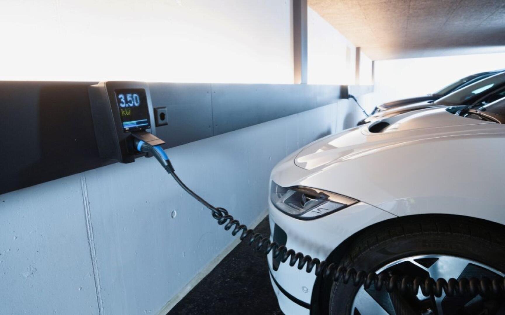 Ein weißes Elektroauto ist an einer Wandladestation in einem Parkhaus angeschlossen. Die Anzeige am Ladegerät zeigt 3,50 kWh an. Auf den angrenzenden Plätzen sind weitere Elektrofahrzeuge geparkt.
