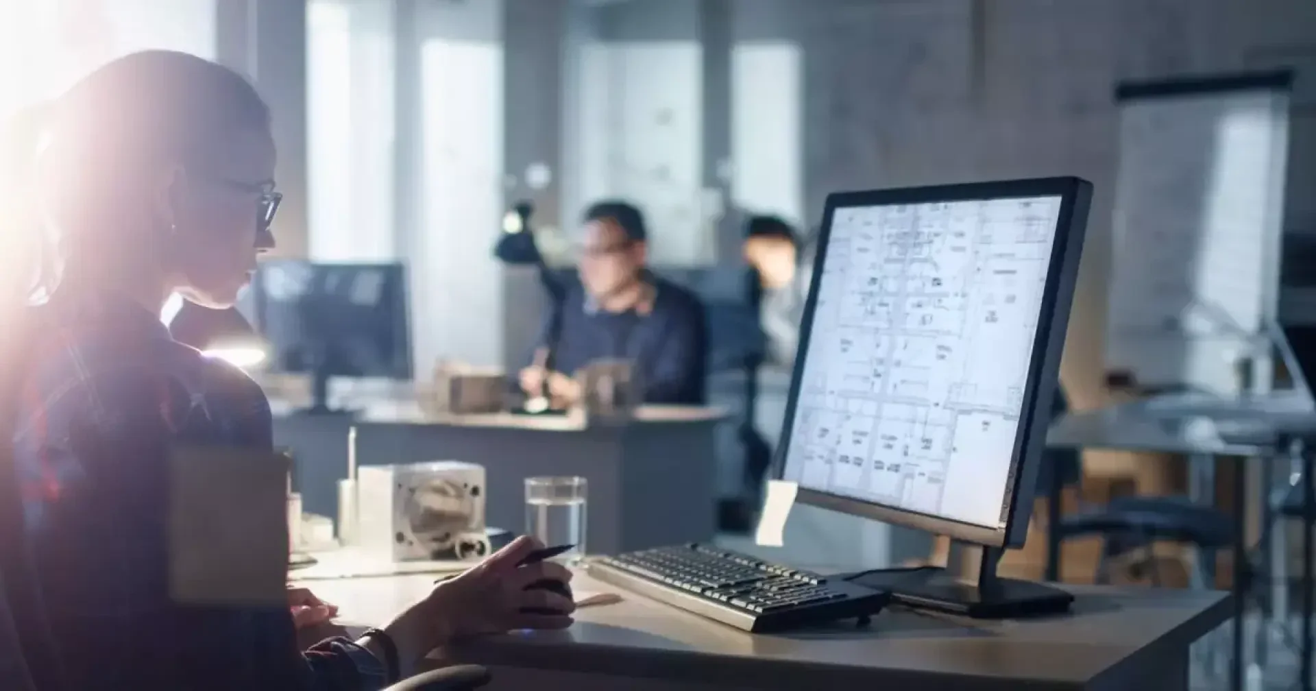 Une personne travaillant à un bureau dans une pièce faiblement éclairée et regardant des projets architecturaux ou des dessins techniques sur un grand écran d'ordinateur, avec les employés en arrière-plan.
