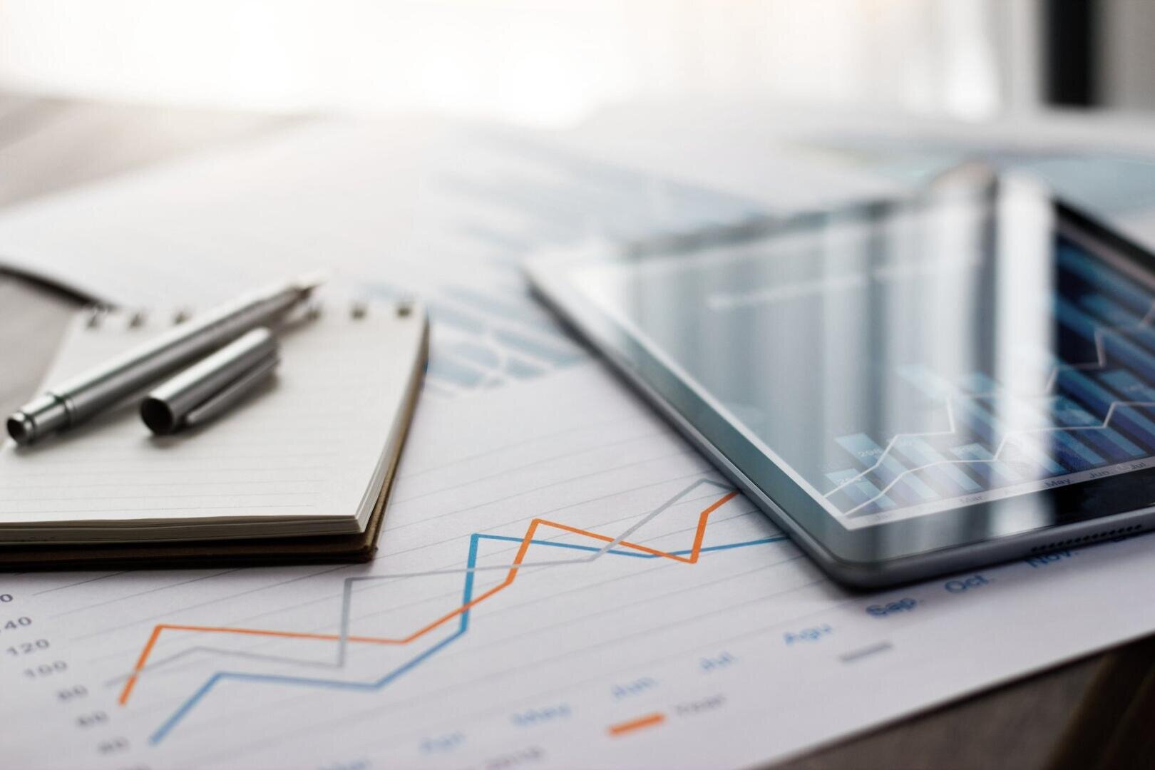 Gros plan sur un bloc-notes avec des stylos, des diagrammes en papier avec des lignes de données et une tablette avec un diagramme, tous disposés sur un bureau avec de la lumière naturelle.
