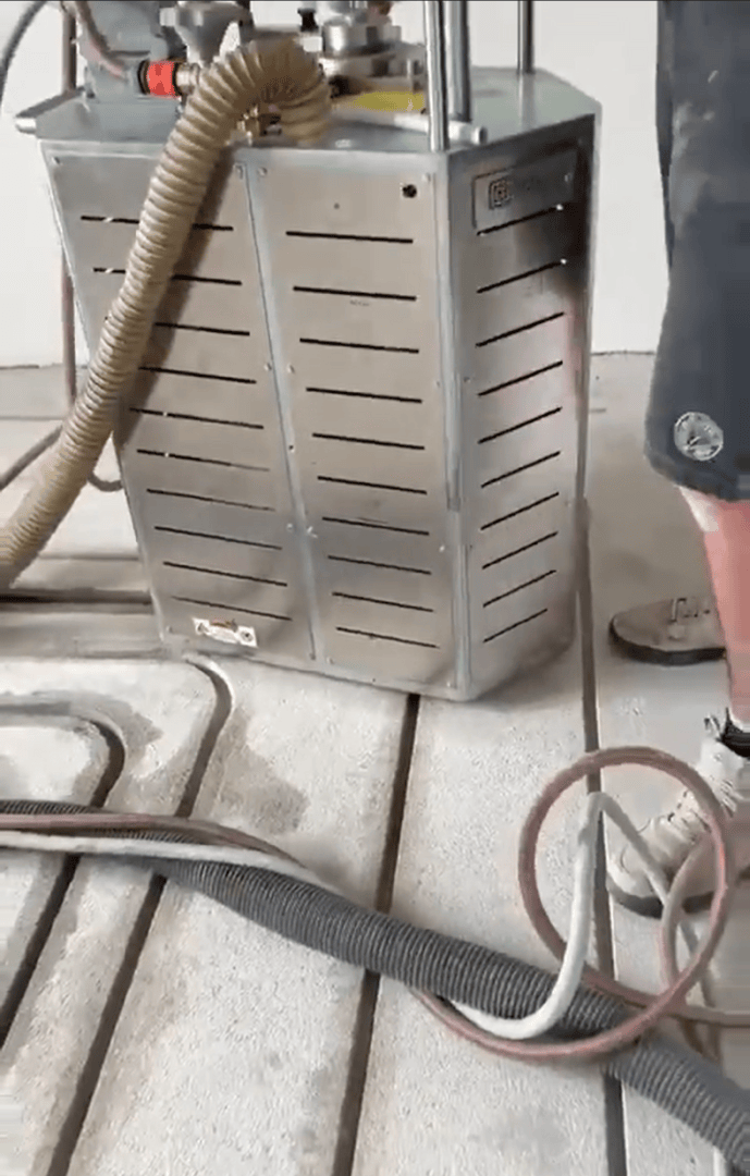 Eine Person in kurzen Hosen und Turnschuhen steht neben einer großen Industriemaschine aus Metall mit Entlüftungsöffnungen, umgeben von mehreren dicken Schläuchen, die auf einem Betonboden liegen.