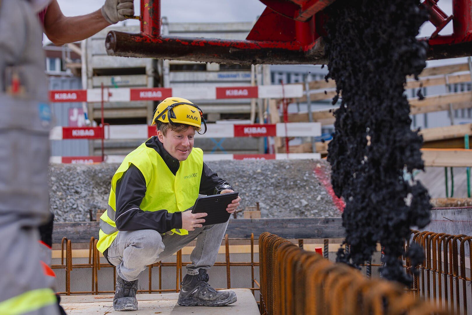 Ein Bauarbeiter, der einen gelben Helm und eine Sicherheitsweste trägt, hockt mit einem Tablet und beobachtet, wie auf einer Baustelle Asphalt gegossen wird, während im Hintergrund Betonstahl und Baumaterialien zu sehen sind.