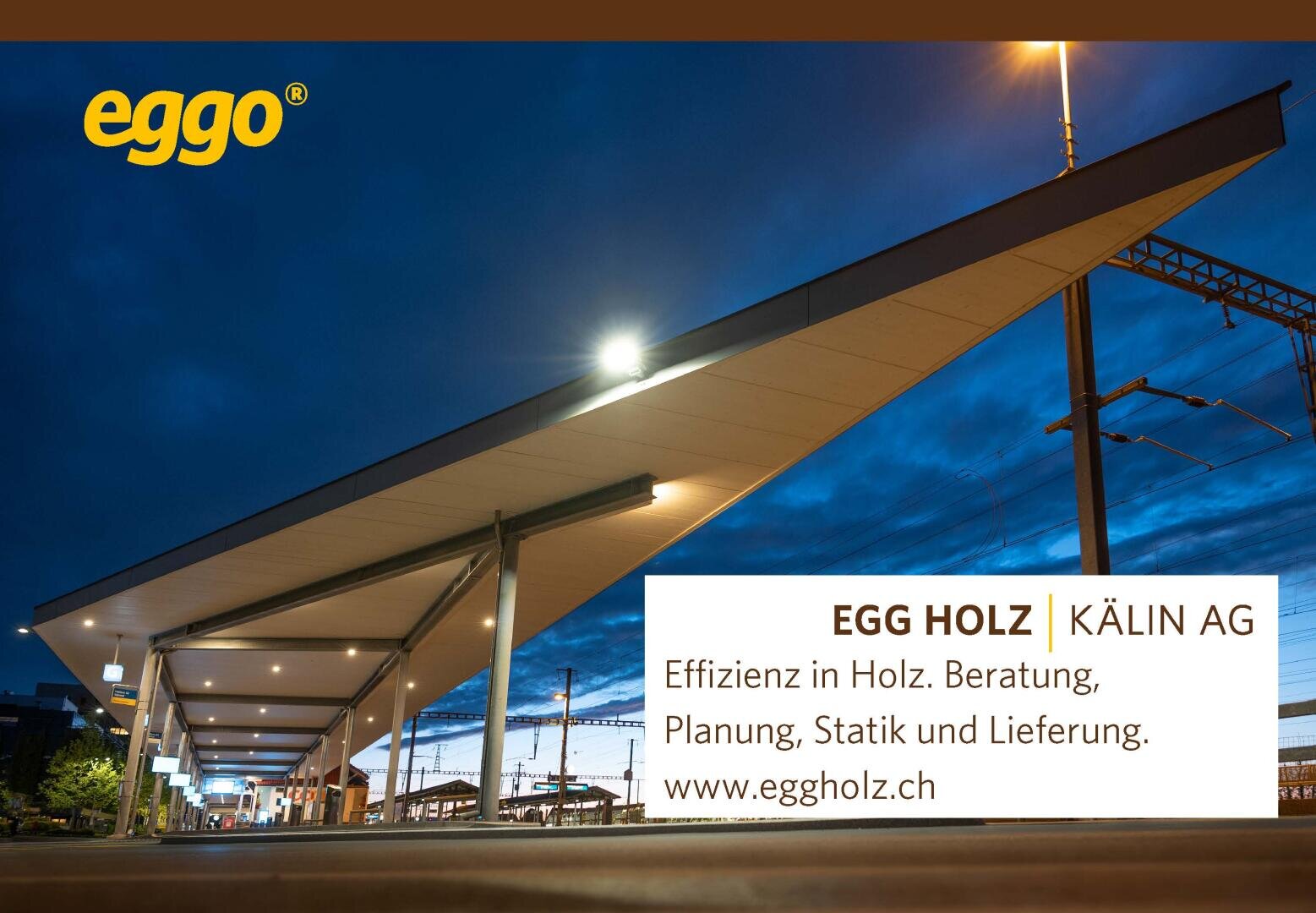 Un quai moderne, bien éclairé, avec un long auvent au crépuscule. Le ciel est d'un bleu profond et des lignes électriques sont visibles. Au premier plan, on peut voir une publicité de EGG HOLZ | KÄLIN AG avec des informations de contact.