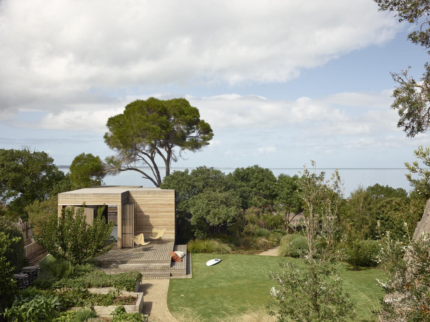 Ein modernes Holzhaus mit Terrasse liegt in einem üppigen grünen Garten mit Blick auf das Meer; ein großer Baum und verschiedene Pflanzen umgeben das Haus unter einem teilweise bewölkten Himmel.