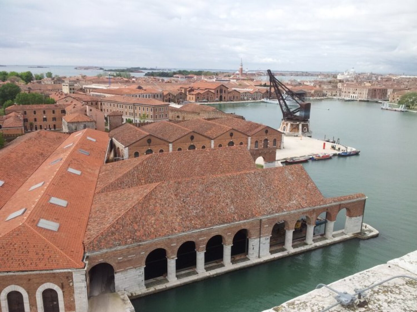 Ein Blick auf das Arsenale von Venedig. Man sieht historische Backsteingebäude mit roten Ziegeldächern neben einem Kanal, eine alte Krankonstruktion und Boote, die unter einem bewölkten Himmel in der Nähe der Uferpromenade anlegen.