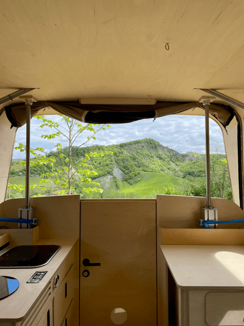 Blick aus dem Inneren eines Wohnmobils durch eine offene Hintertür auf einen üppig grünen Hügel unter einem teilweise bewölkten Himmel, mit einer kleinen Küche und Arbeitsplatte im Vordergrund.