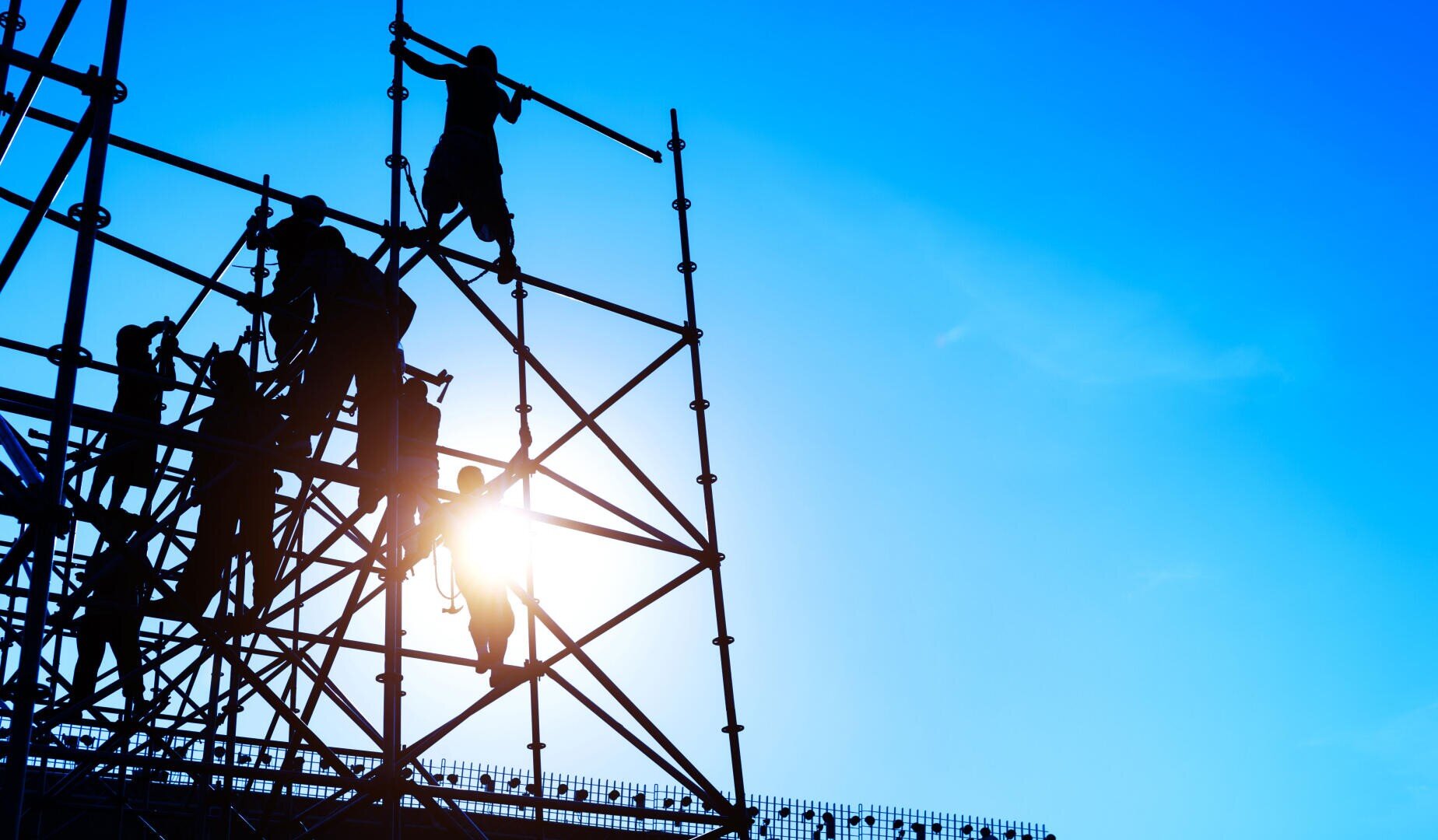 Silhouettierte Bauarbeiter klettern auf ein hohes Metallgerüst und montieren es vor einem strahlend blauen Himmel, wobei die Sonne durch die Struktur scheint.