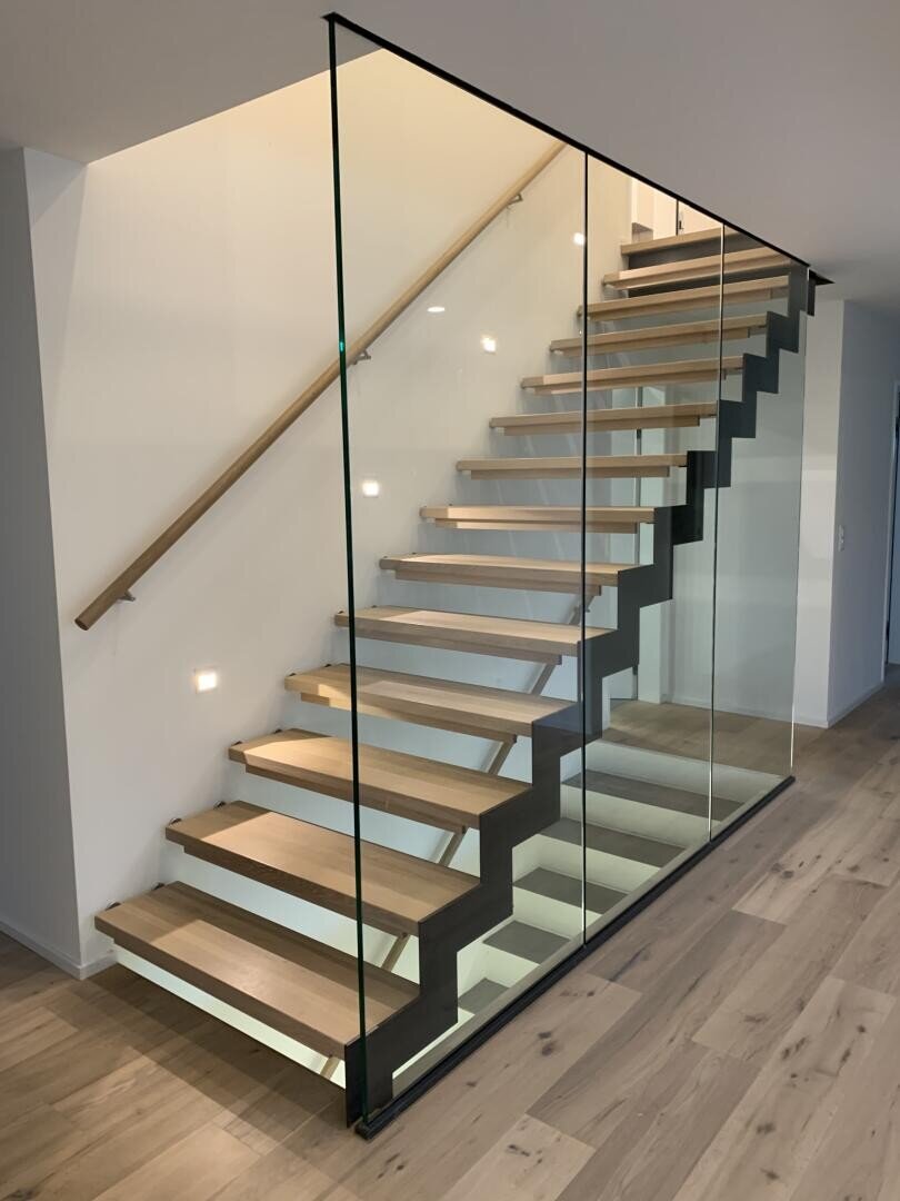 Un escalier moderne avec des marches en bois flottantes, un cadre métallique noir et des panneaux latéraux en verre transparent, intégré dans un intérieur minimaliste avec un sol en bois clair et des murs blancs.