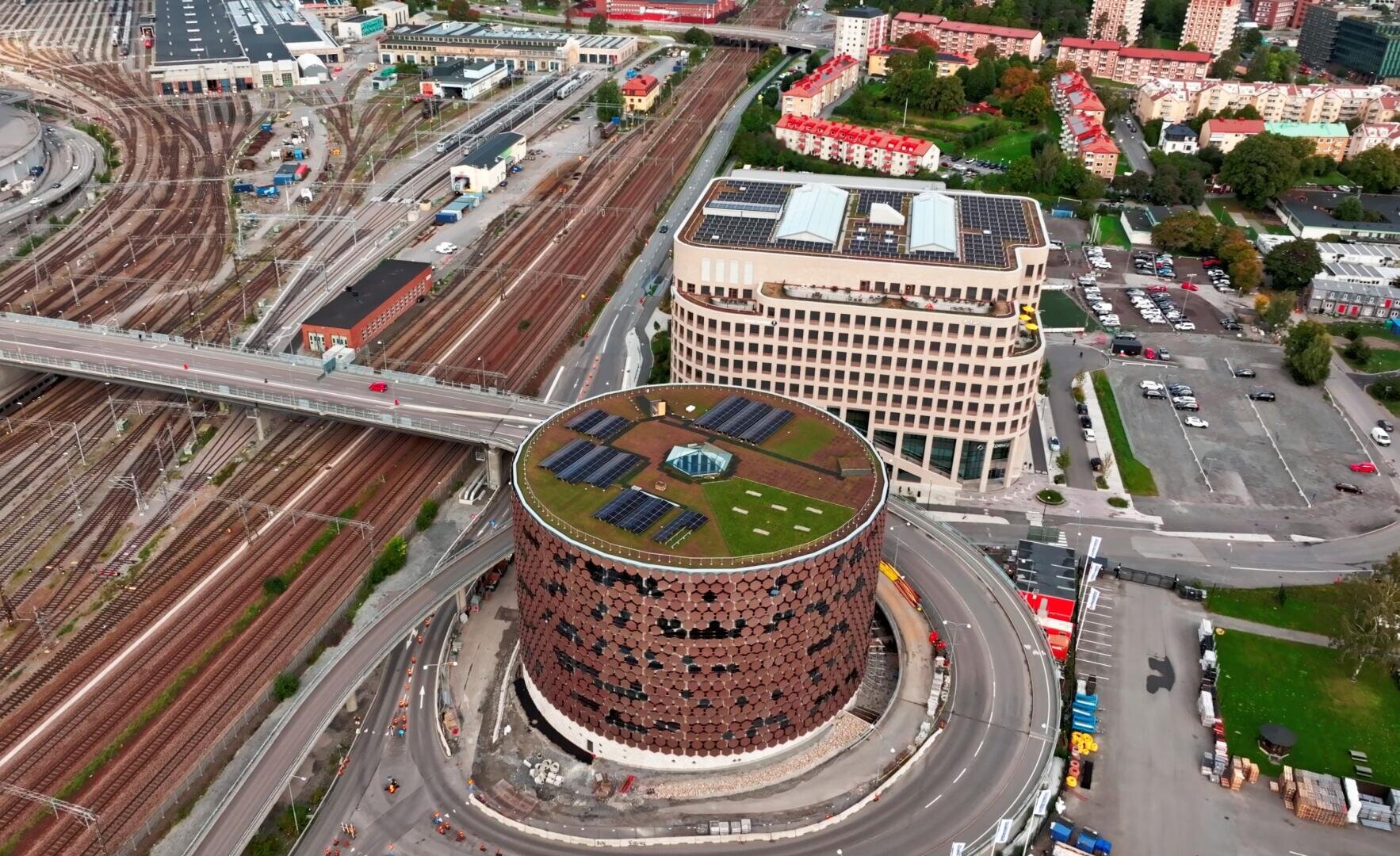 Luftaufnahme eines runden Gebäudes mit begrüntem Dach und Sonnenkollektoren, neben Bahngleisen und einem modernen mehrstöckigen Gebäude, umgeben von Straßen und nahe gelegenen Wohn- und Gewerbegebieten.