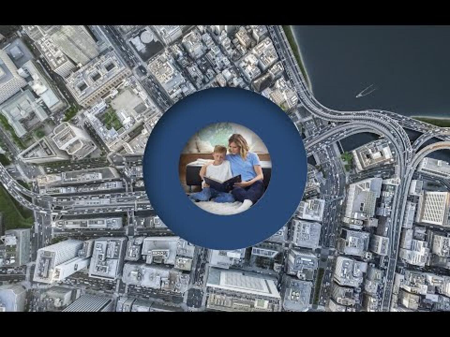 Luftaufnahme einer Stadt mit Straßen und Gebäuden, mit einem kreisförmigen Einschub in der Mitte, der eine Frau und ein Kind zeigt, die gemeinsam auf einer Couch ein Buch lesen.