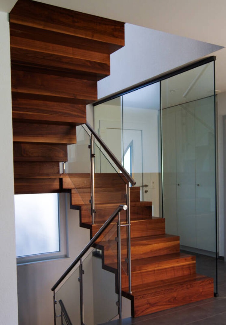 Eine moderne Holztreppe mit Metallgeländer im eckigen Design. Glaspaneele säumen eine Seite und verleihen ihr einen eleganten, modernen Touch. Die umgebenden Wände sind weiß, und ein Fenster lässt Tageslicht herein.