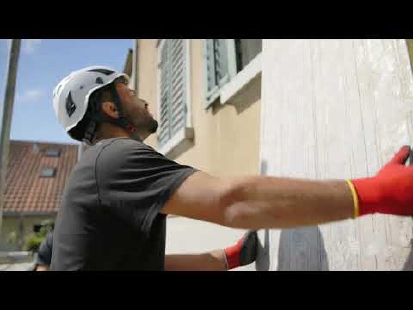 Ein Mann mit weißem Schutzhelm und roten Handschuhen befestigt ein großes Holzbrett an einem Fenster eines beigen Hauses und bereitet sich möglicherweise auf einen Sturm vor. Im Hintergrund sind die Dächer und der blaue Himmel zu sehen.
