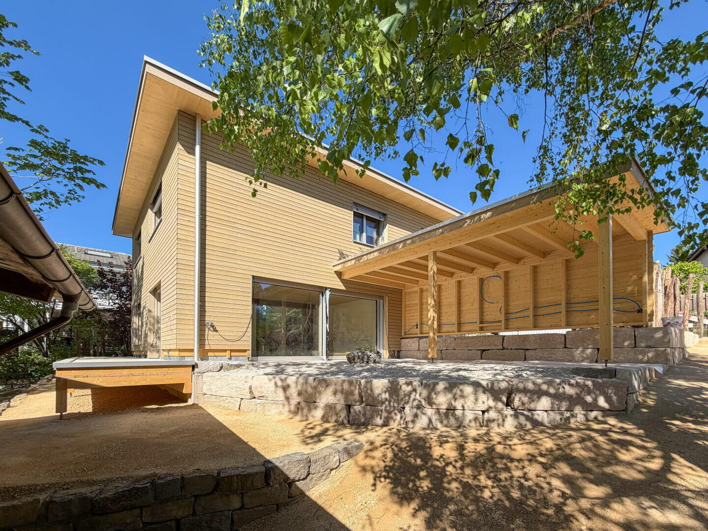 Ein modernes zweistöckiges Holzhaus mit großen Fenstern und einer überdachten Terrasse, umgeben von Bäumen und auf einem erhöhten Steinfundament unter strahlend blauem Himmel.